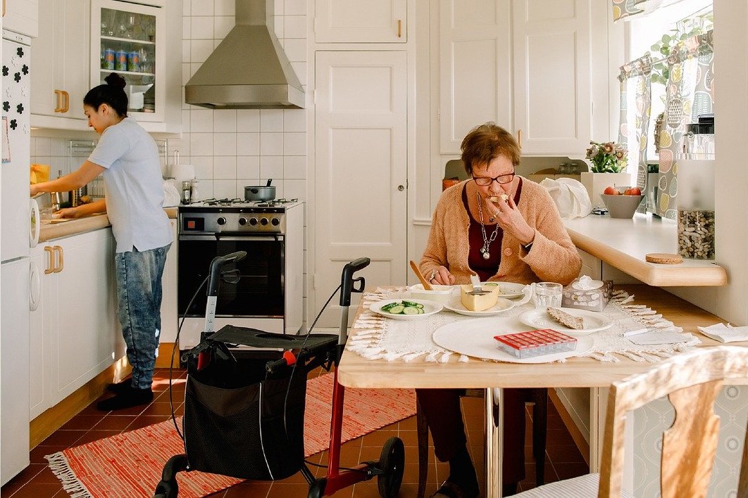 Foto av eldre dame som sitter på kjøkkenet, med hjelpearbeider i bakgrunnen.