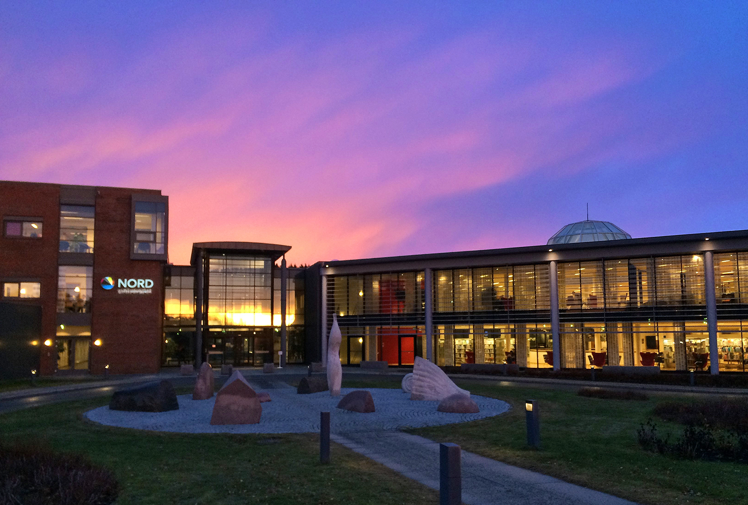 Nords fasade med solnedgangsfarger på himmelen bak. Man ser hovedinngangen i glass, med murbygget med Nords logo på venstre side og glassbygningen med kuppel som huser biblioteket på høyre side. Plenen foran bygningen er grønn. En gnagsti leder opp til hovedinngangenog krysser en liten rundt plann med steinskulpturer som til dels har abstrakte former og til dels er skjellformede.