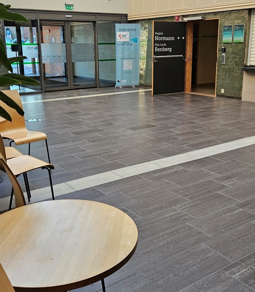 An fairly big area with a lot of empty floor. There are some chairs and tables on the left hand side in the foreground. In the background, there are two sets of sliding doors leading outside. In side and to the right of these sliding doors, there is an open door on which it says "Regine Normann" and "Elsa Laula Renberg", inside this door there is a small space where parts of a door and coat hangers are visible on the left wall.