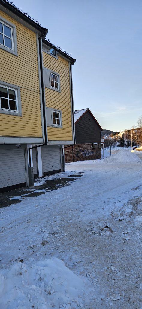 Snow-covered pavement with a yellow wooden three-floor apartment building on its left hand side. There seems to be a gap between this building and the next one, which is a lower brown, wooden building with a red-brick foundation and a pitched roof.