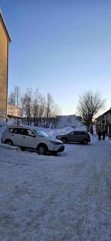 A walking path with a yellow wooden building with a small parking space in front on the left side and a low and long brown building on the right side. At the end of the path there's another building.