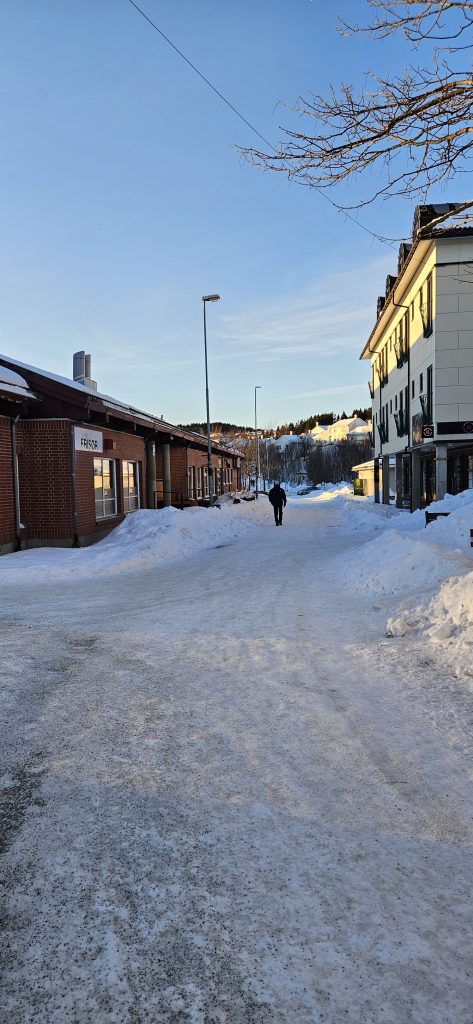 A walking path between two rows of buildings with various signs on them. At the end of the path, there's a car on a road below a small hill.