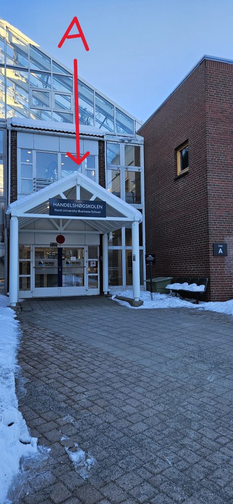 An entrance in glass and white metal with the sign "Handelshøgskolen - Nord University Business School" above it. The entrance is labelled "A" in the pciture. To the right of the entrance, the wall is of red brick, and there is a small sign on the wall where "A" is written.