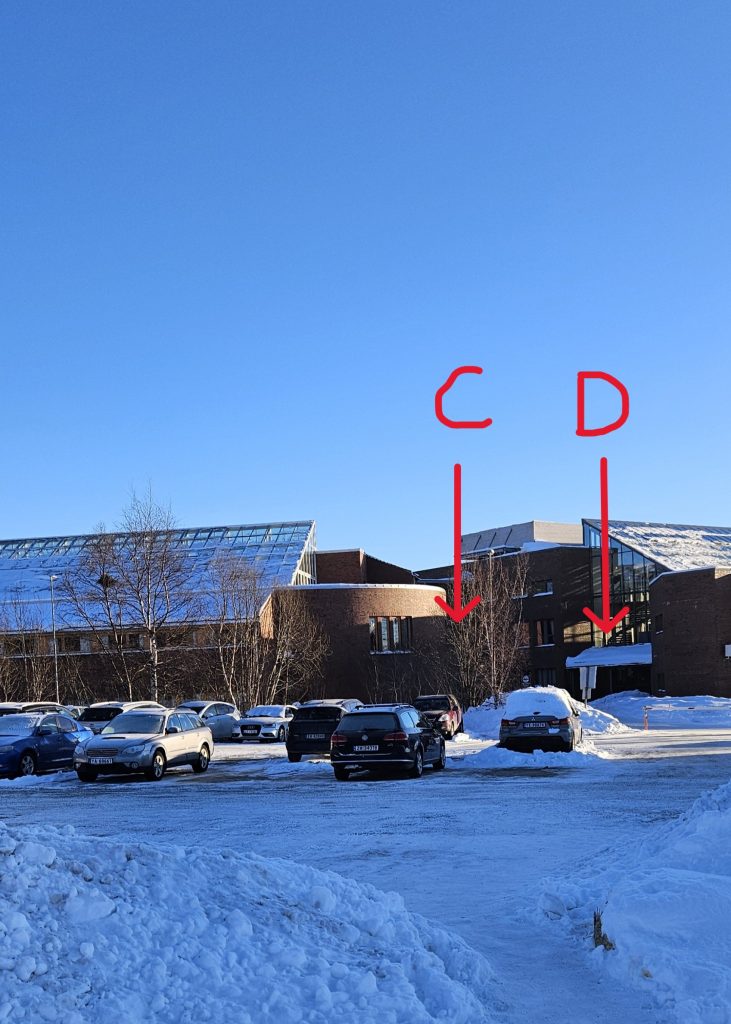 A small parking lot in front of the main building of the university, a red brick building with slanting glass roofs. There is a small gap between two parts of the building. The entrance to the leftmost part is labelled "C" in the picture, whereas the entrance to the rightmost part is labelled "D".