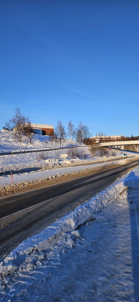 A strip of pavement next to a road which has a walking path on the other side. Between the road and the walking path, there is a sign pointing across the road where it says "Mørkvedlia studentboliger". A bit further down there is a metal walking bridge across the road, which starts at the walking path. On the hill above the walking path there is a big building. Another big building can be seen in the backgroun further down the road after the bridge.