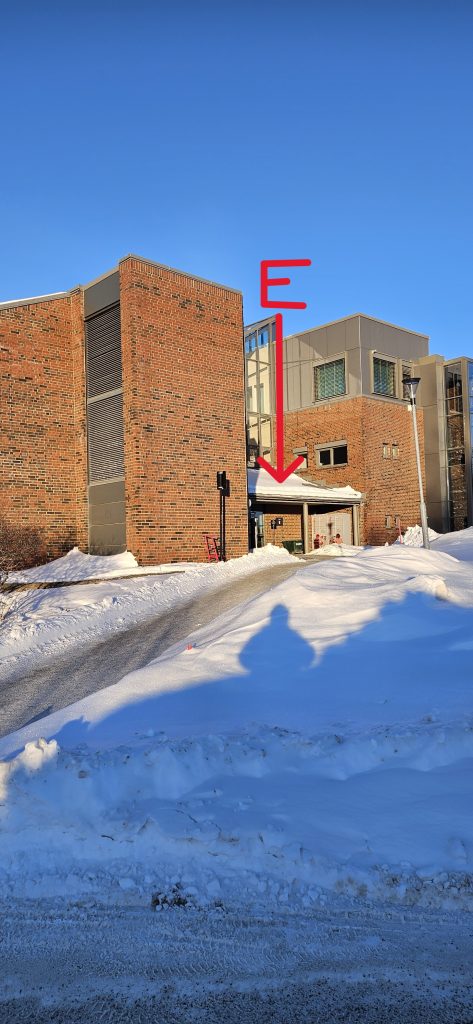 A path up a small slope leading to a the entrance of a red brick building. The entrance is labelled "E" in the picture and has a slanting glass roof directly above it.