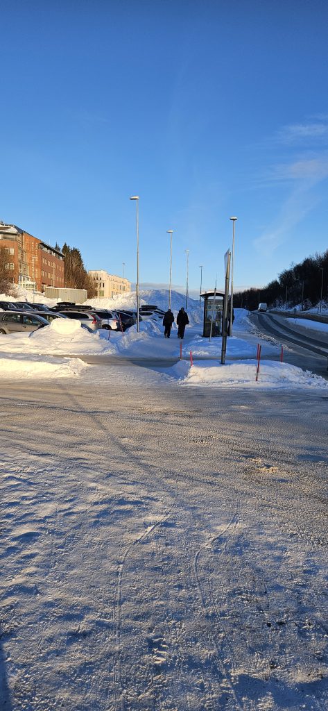 A crossing over to a pavement with a bus stop. On the left of the pavement there is a parking lot, and there are a few buildings on the other side of the parking lot. On the right of the pavement there is a road.