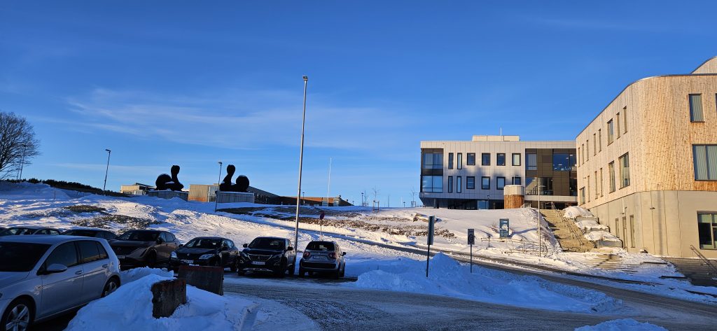 A parking lot to the left and a couple of buildings to the right of a small rpad going up a hill. On top of the hill are two dark wooden sculptures, and the roof of a red bruck building can be seen behind them.