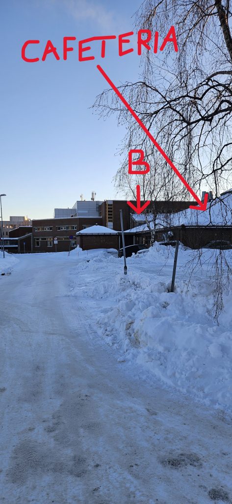A pathway in the snow towards a couple of red brick buildings. The closest one is labelled "Cafeteria" in the picture, whereas the entrance of the building which is partly behind it is labelled "B".